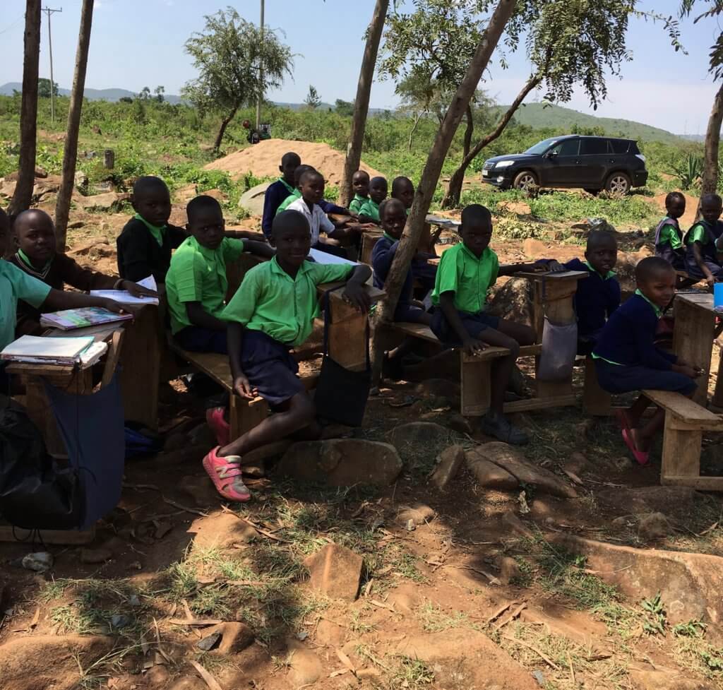 students in Kenya sitting at rough hewn desks under a tree. There is no fence, so securing the perimeter is needed here.