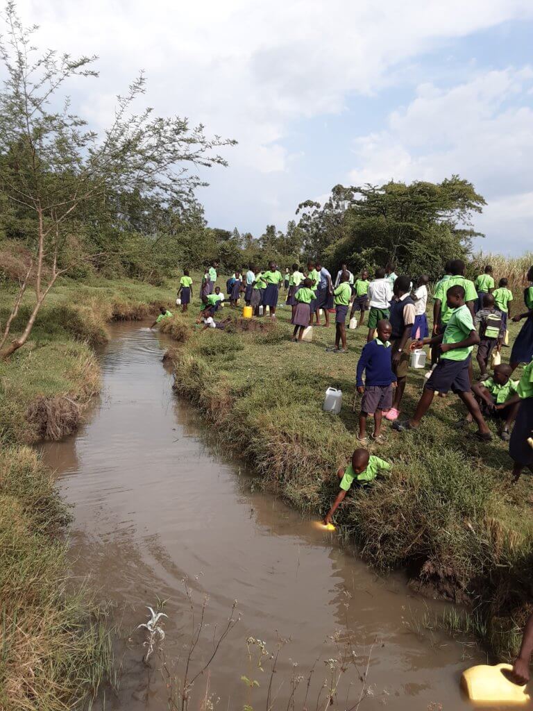 Many young children standing at a muddy riverbank collecting water in yellow jerry cans